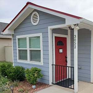 Small gray house with white trim and a red front door. The number 3052 is on the porch post. There are two windows with green blinds, and a small garden with green plants in the front.