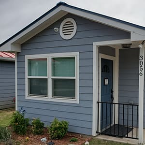 A small blue house with white trim, featuring a front porch with a black railing, double windows and the number 3056 on the right side.