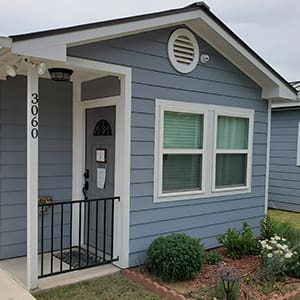 A small gray house with white trim, two windows, and a covered entrance with a black railing. The address number "3060" is displayed vertically near the door. Bushes and plants adorn the front yard.