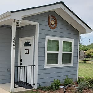 A small, light blue house with white trim, featuring a front door with the number 3064 and a wooden emblem above the window. The house has a simple garden and a grass lawn.