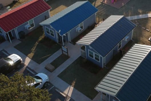 Aerial view of a small cluster of single-story houses with red, blue, and gray roofs. The houses are arranged along a pathway with parked cars visible nearby.