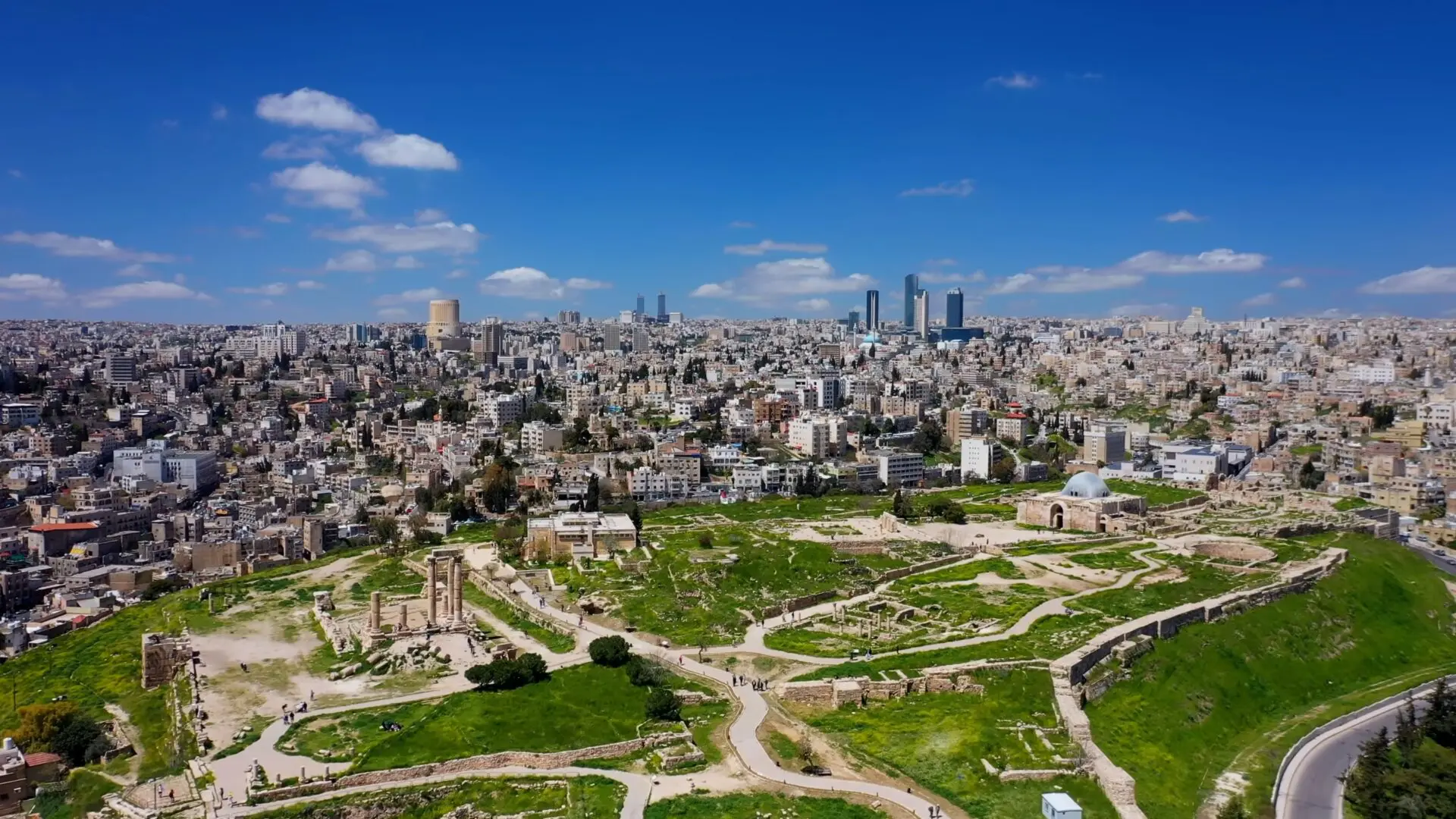 Amman Jordan wallpaper Aerial view of a cityscape with a mix of modern and historical buildings, lush green areas, and ruins in the foreground under a clear blue sky with scattered clouds.