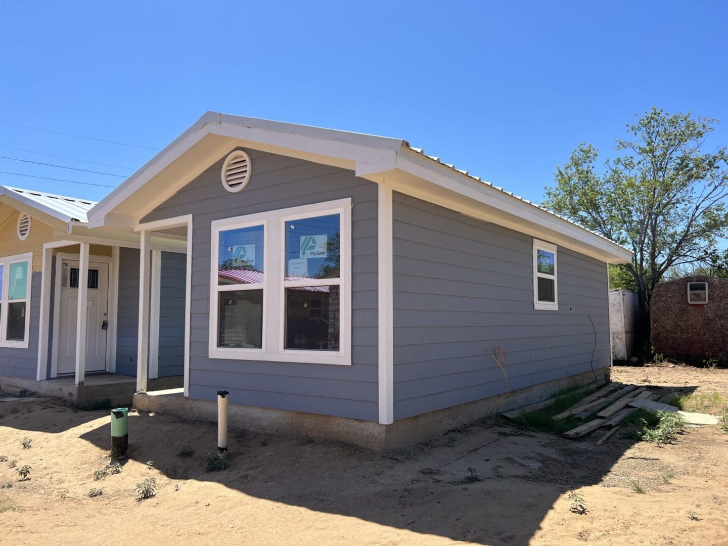 A small, grey single-story house with a gable roof, white trim, and two front-facing windows. It is set on a dirt plot with minimal vegetation and a tree in the background.