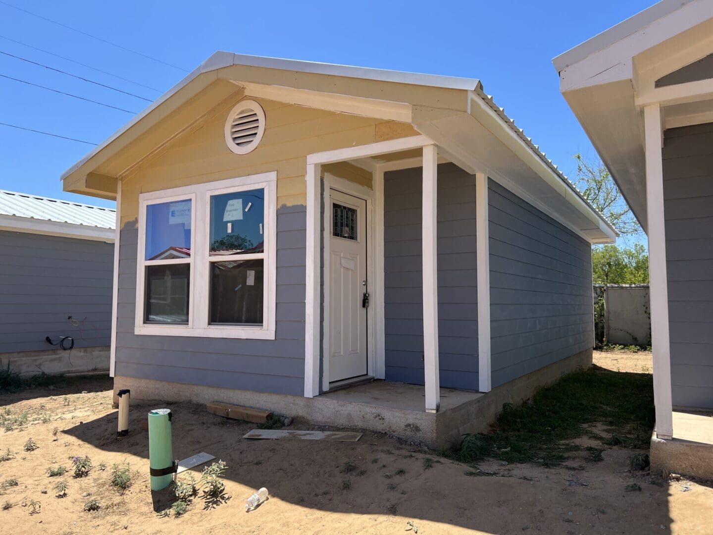 A small single-story house with a yellow and gray exterior, white trim, and a front door facing forward. The ground surrounding the house is bare and appears to have some patches of grass and construction debris.