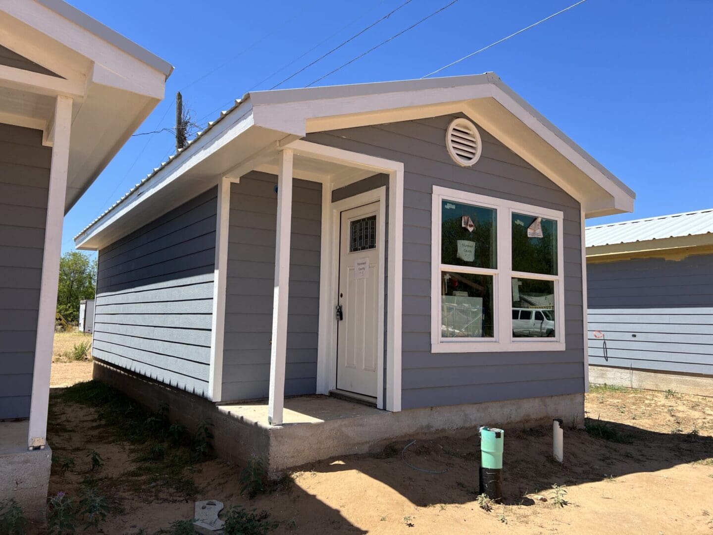 A small, single-story gray house with a white front door and porch, under a clear blue sky. Construction materials are visible around the foundation.