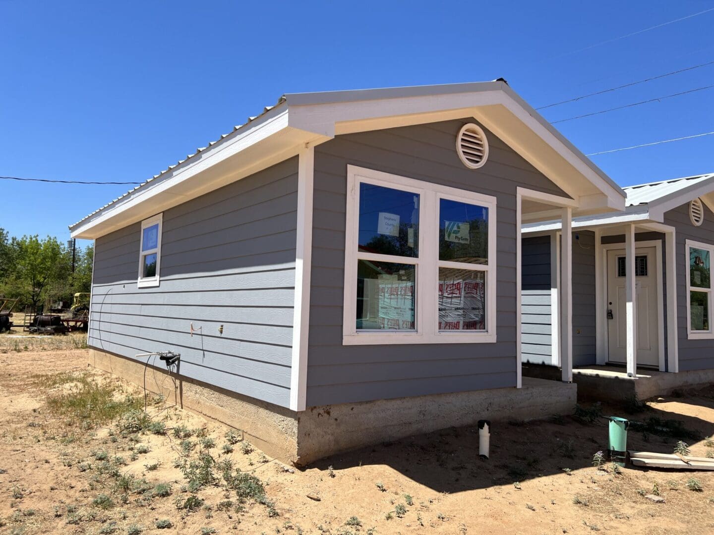 A small gray house with a white roof and white-framed windows sits on a dirt lot under a clear blue sky. The house is elevated on a concrete foundation with some building materials nearby.