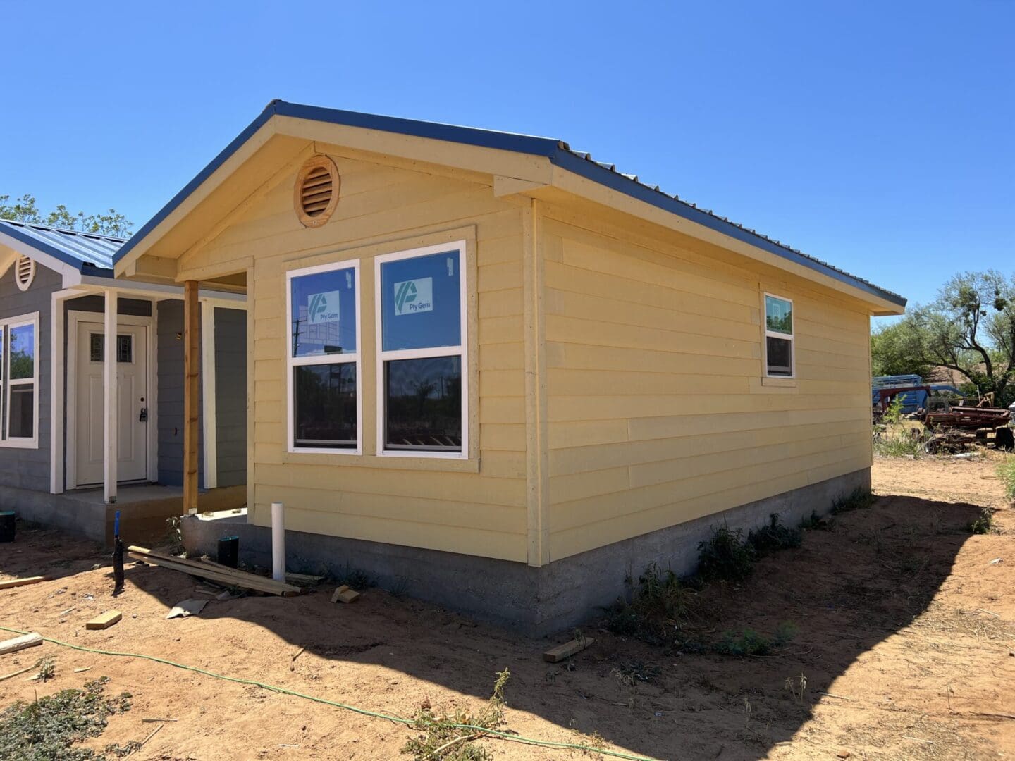 A single-story yellow house with two front windows and a small porch is under construction. The surroundings are dry and sparse. The sky is clear and blue.