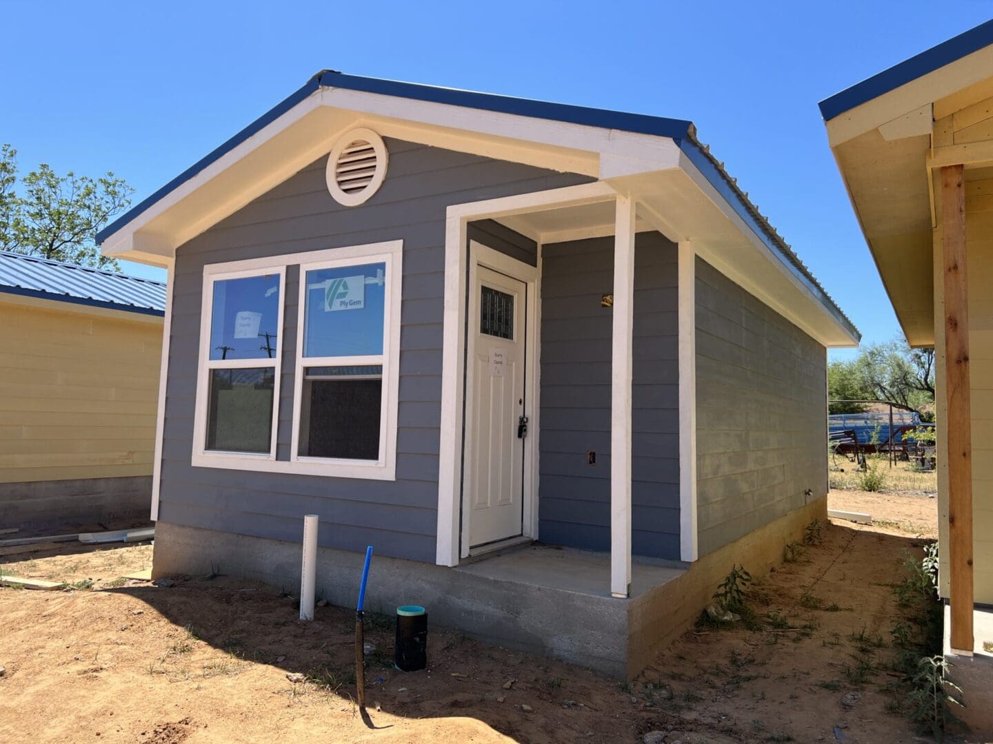 A small gray house with white trim and a blue roof stands on a dirt lot. The house has a front door, two windows, and a small porch area. There's a shovel and a black pipe in the foreground.