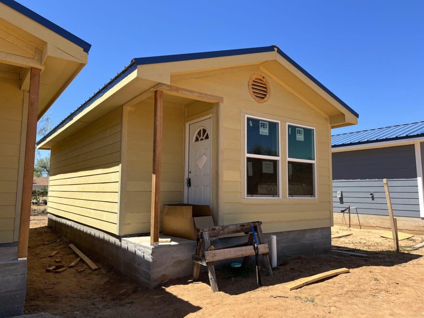A small yellow house under construction with a white door and two windows. Building materials and construction tools are in the foreground.
