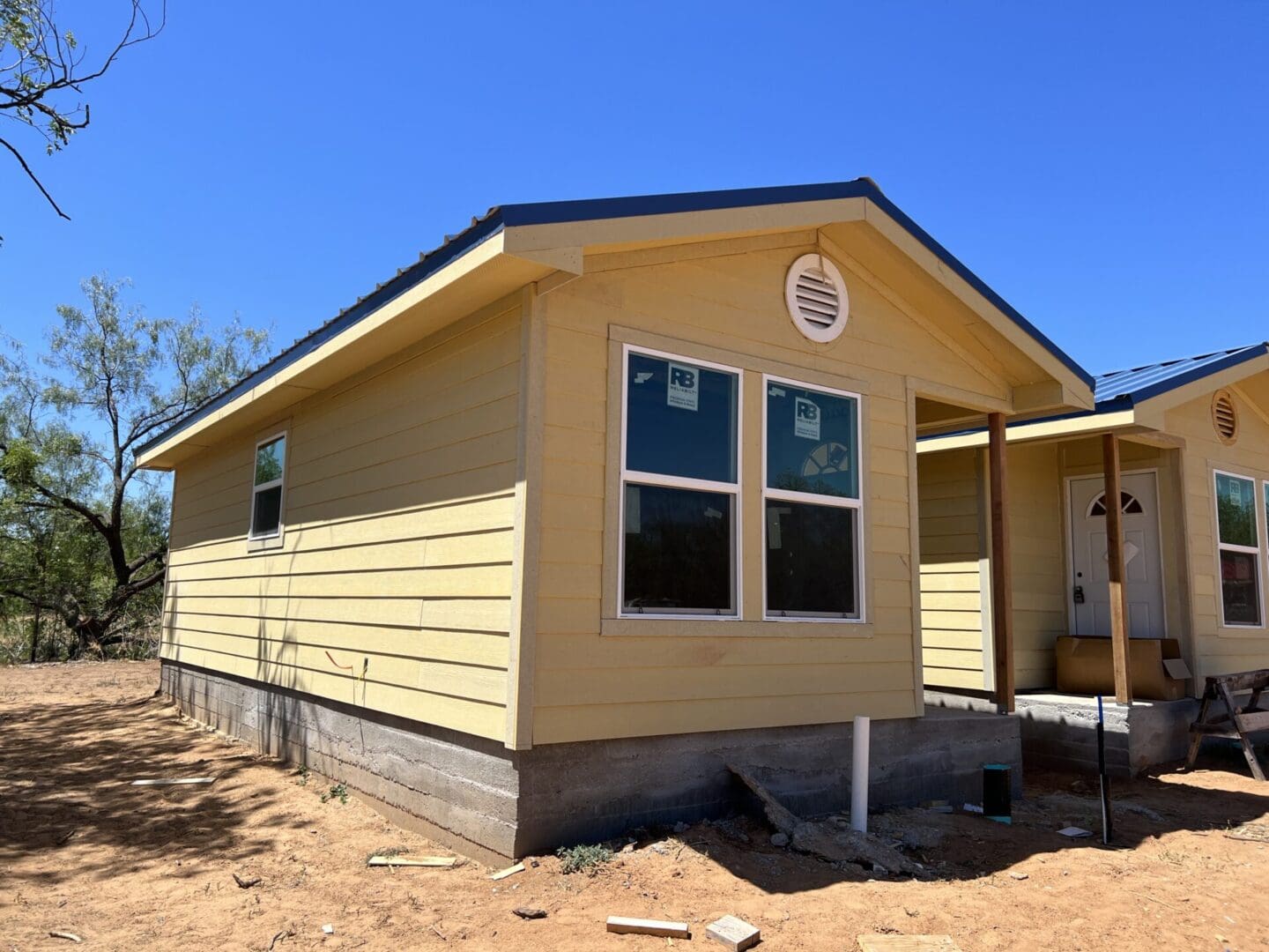 Single-story, yellow house with a blue roof under a clear sky, featuring a front entrance with a small porch and two white-framed windows on the visible side.