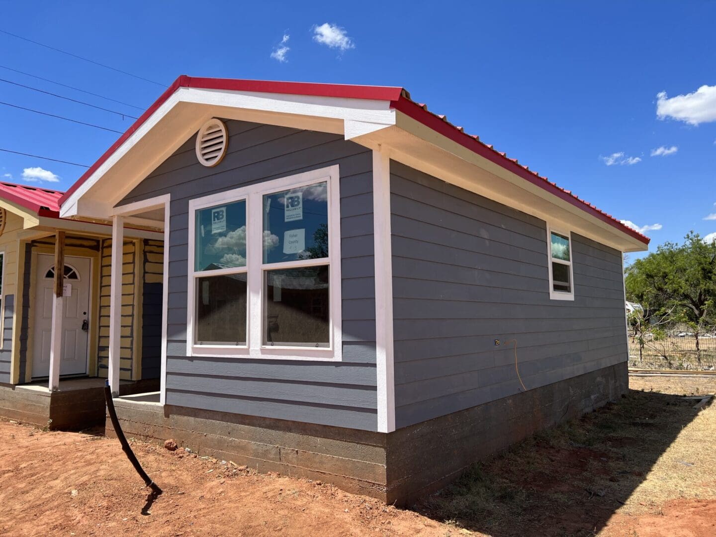 A small grey house with a red roof, two front windows, a circular vent, and a white door, situated on a dirt lot under a clear blue sky with a few clouds.