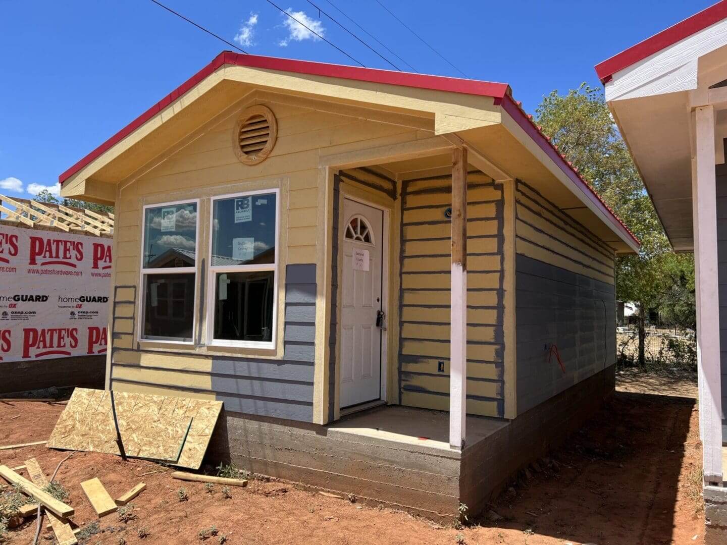 A small, yellow house with brown striped siding, a white door, and a partially installed window stands on a red dirt foundation, with another house and construction materials nearby.