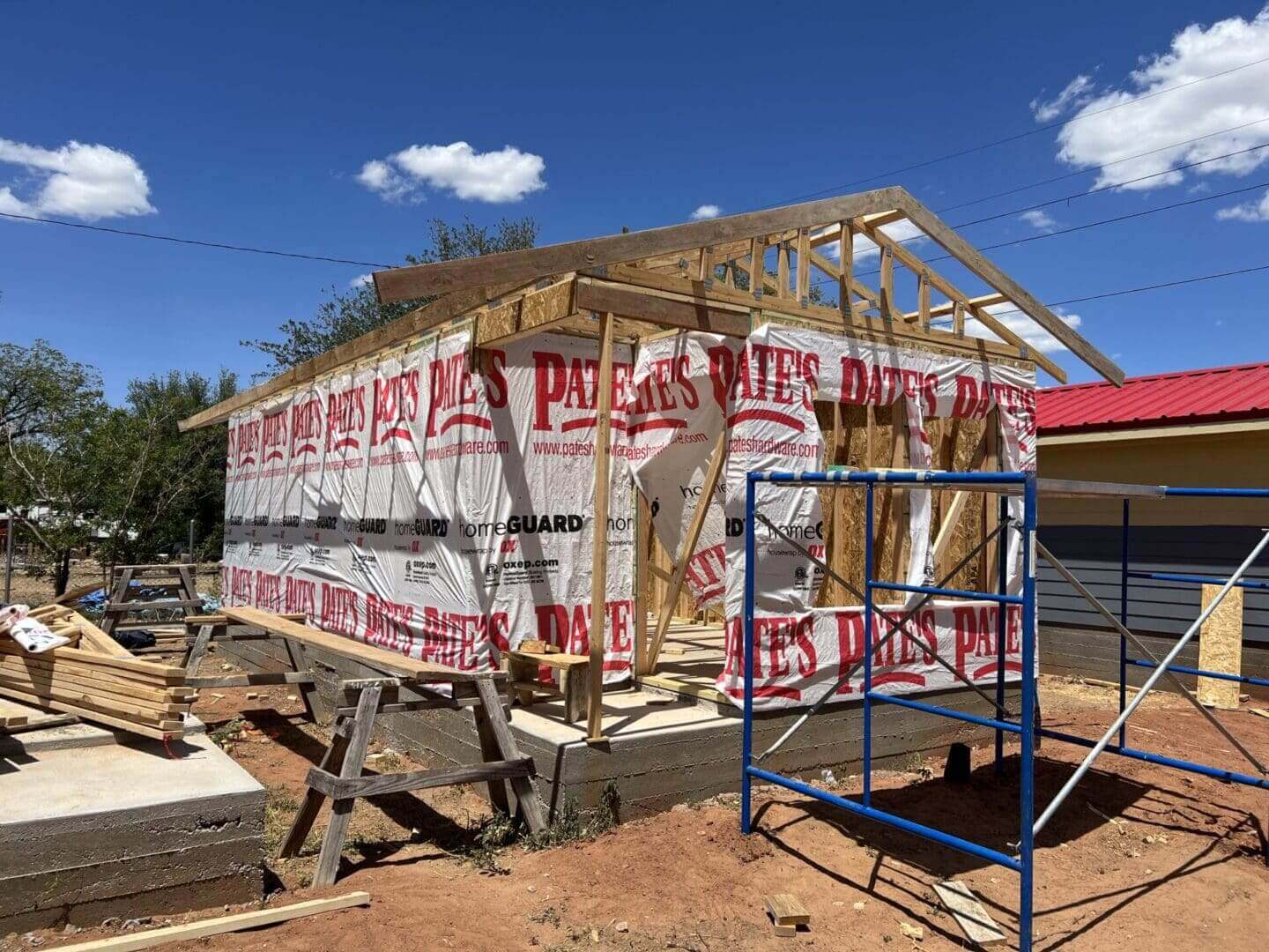 Partially constructed building with wooden framing and protective wrap. A blue scaffolding stands nearby under a clear blue sky.