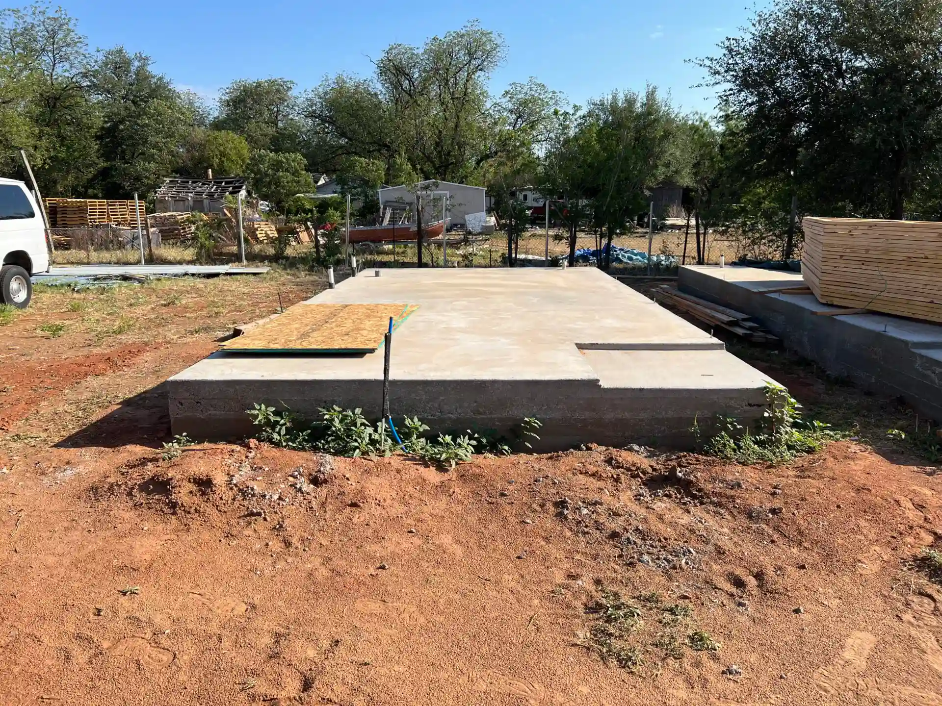 A concrete foundation slab in an outdoor area, with wooden panels and construction materials nearby. Vegetation and a parked vehicle are in the background.