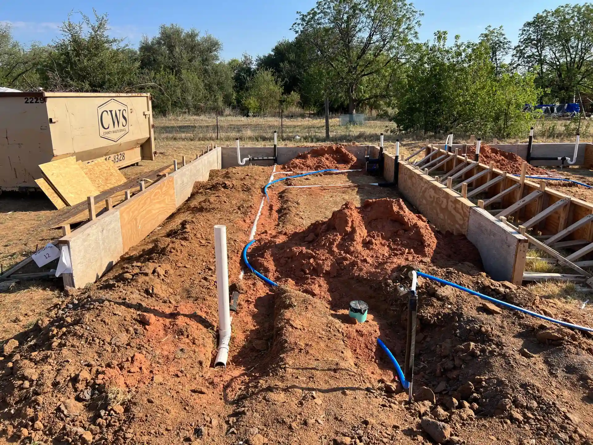 Construction site with foundation partially built, showing a trench with pipes and cables being laid. A large waste container and trees are in the background.