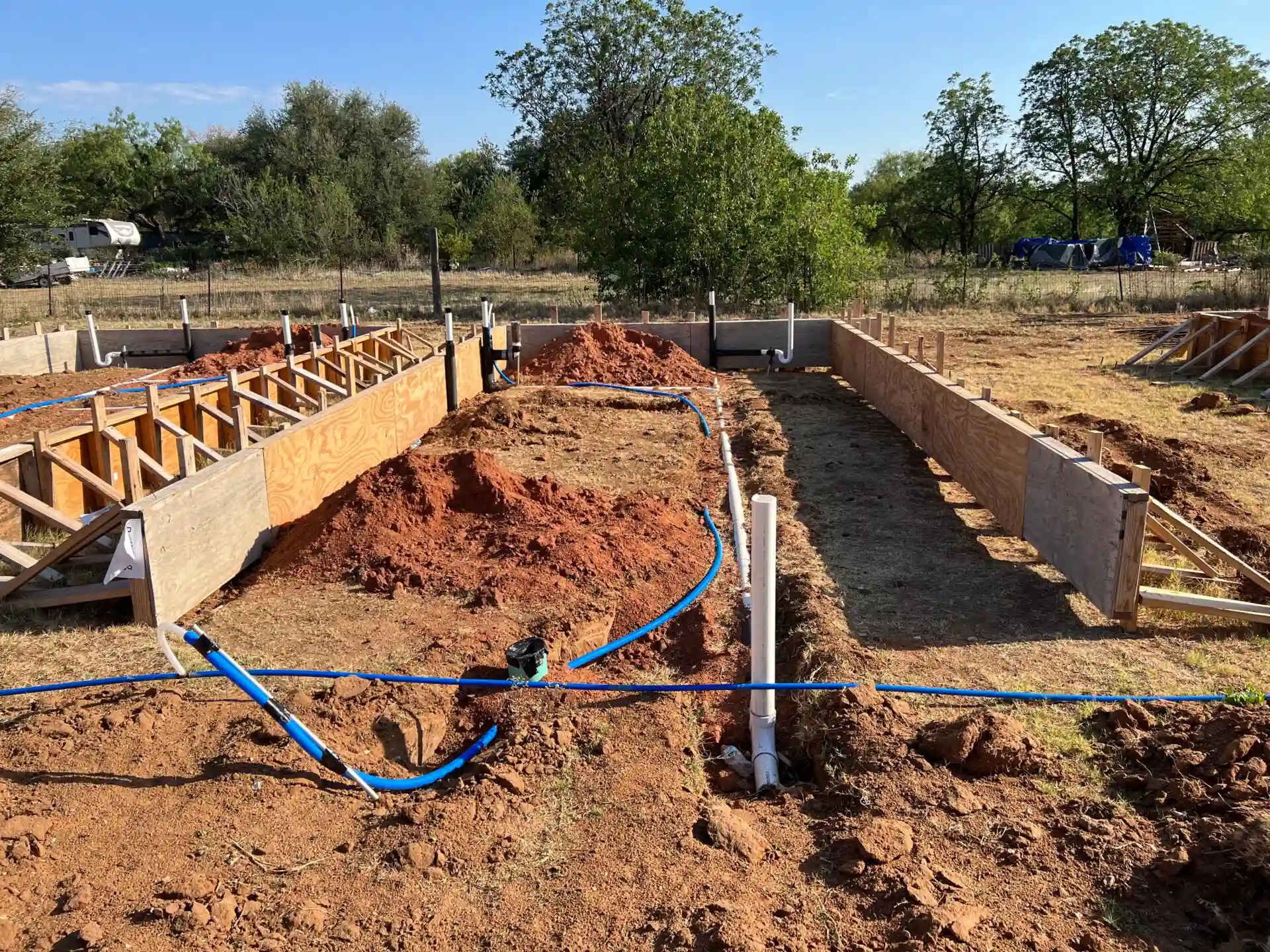 The image shows a construction site with foundation work in progress. Trenches are dug, wooden frames are set, and blue pipes are laid out on the ground. Trees and grass are in the background.