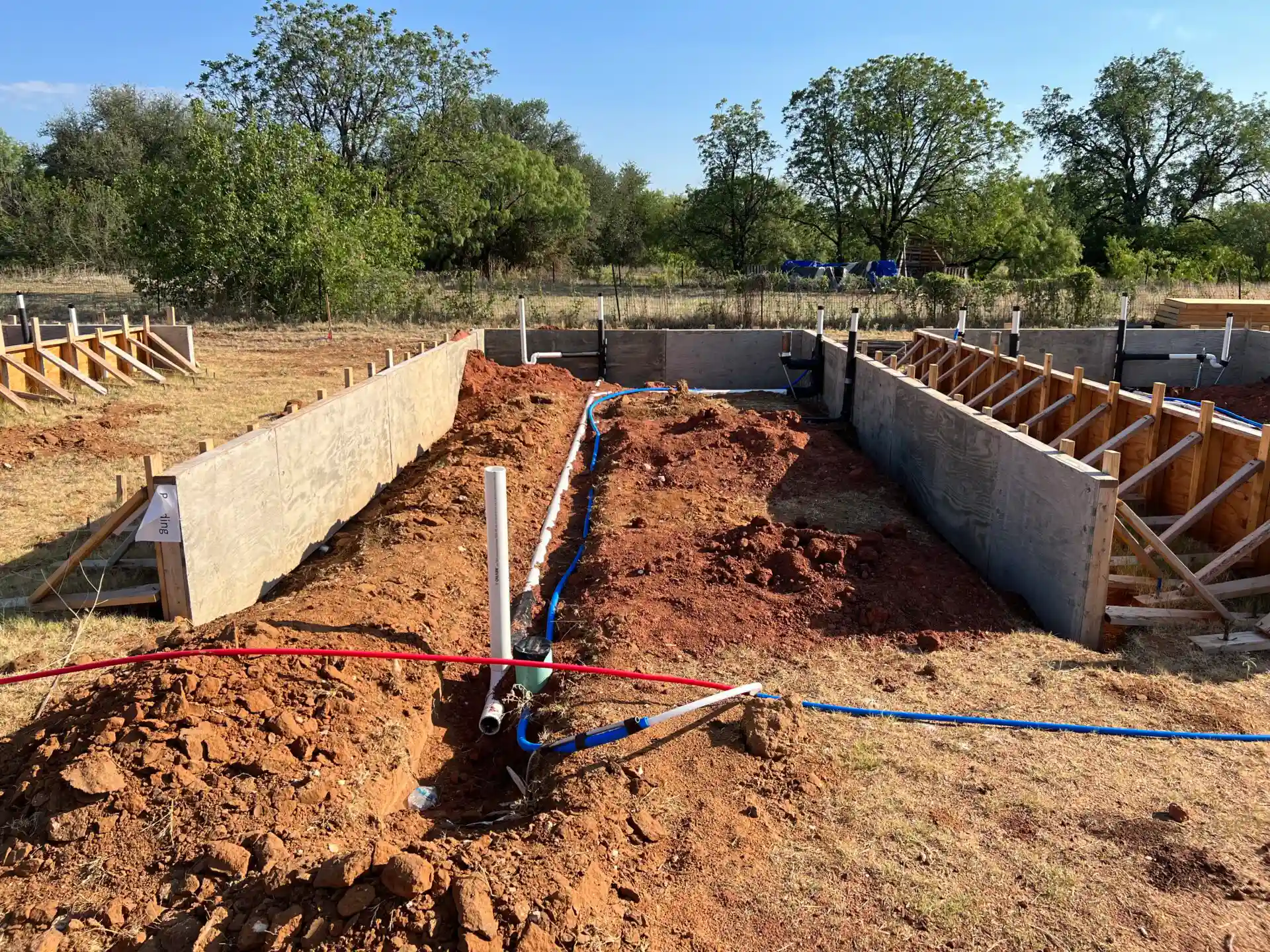 A construction site with foundation trenches filled with dirt and exposed pipes; concrete wall reinforcements and wooden formwork on the sides, amidst trees and grasses in the background.