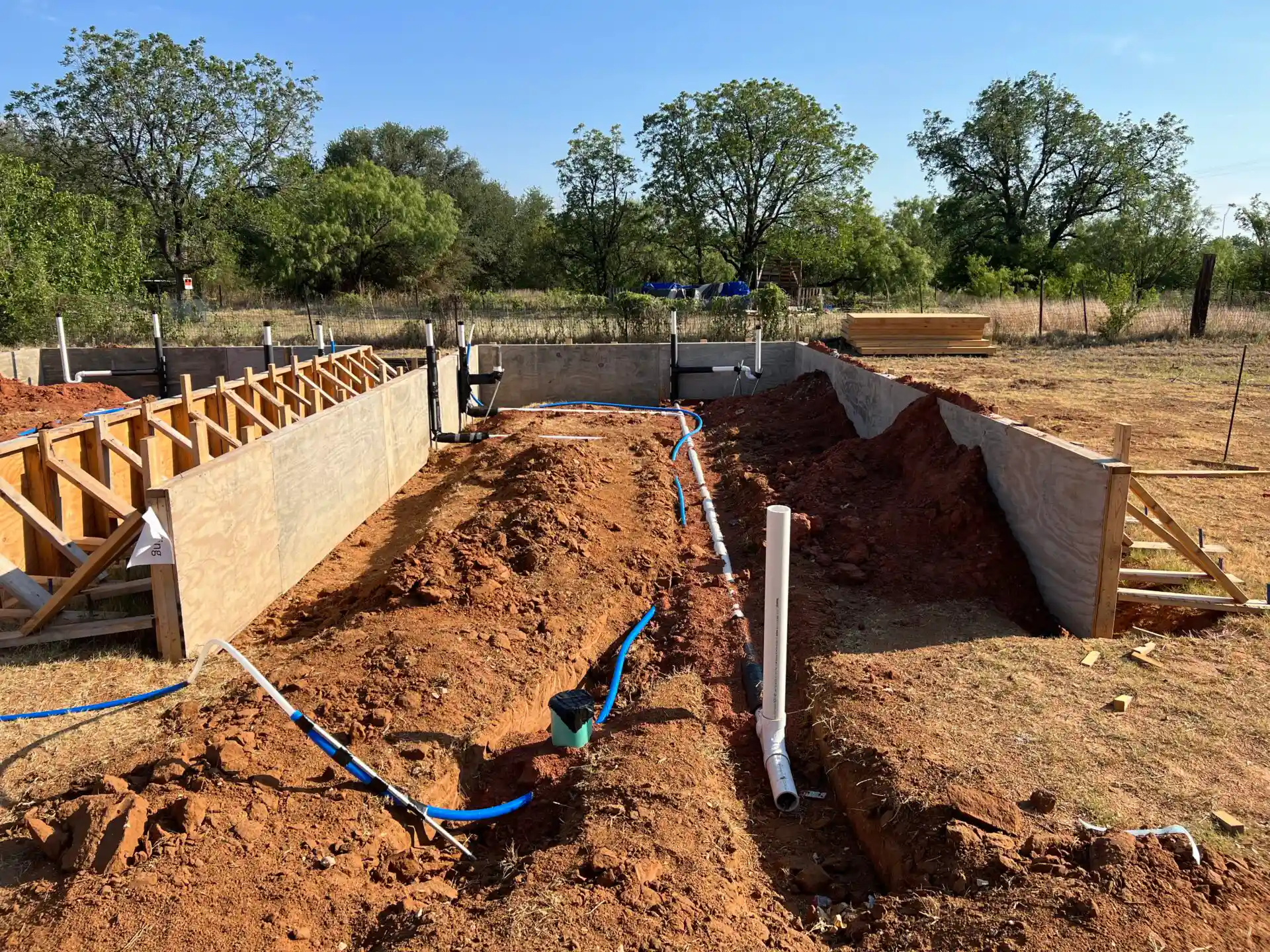 A construction site showing excavated trenches with installed plumbing pipes and partially built foundations surrounded by dirt and trees in the background.