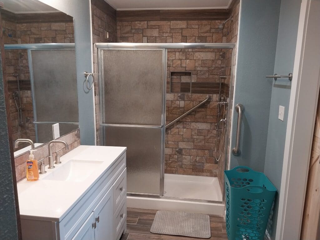 A bathroom featuring a shower with sliding glass doors and a grab bar, a white vanity sink with a mirror, a teal laundry basket, and brick-patterned wall tiles.