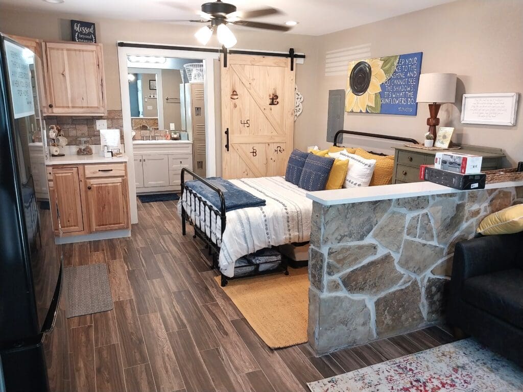 A cozy studio apartment with a bed, stone partition, wooden kitchen cabinets, and a bathroom with a sliding barn door. Decor includes a sunflower painting and modern furnishings.