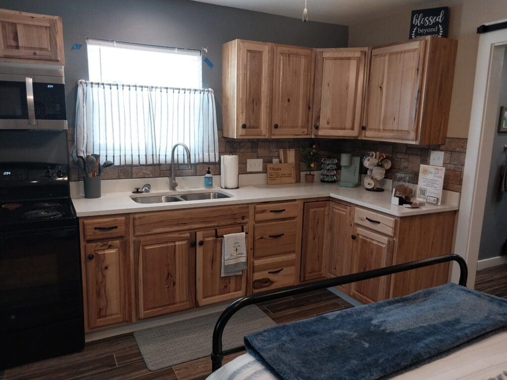 A kitchen layout with wooden cabinets, a stainless steel sink, microwave, and stove. A window with blue curtains is above the sink. There are kitchen items and decor on the countertops.