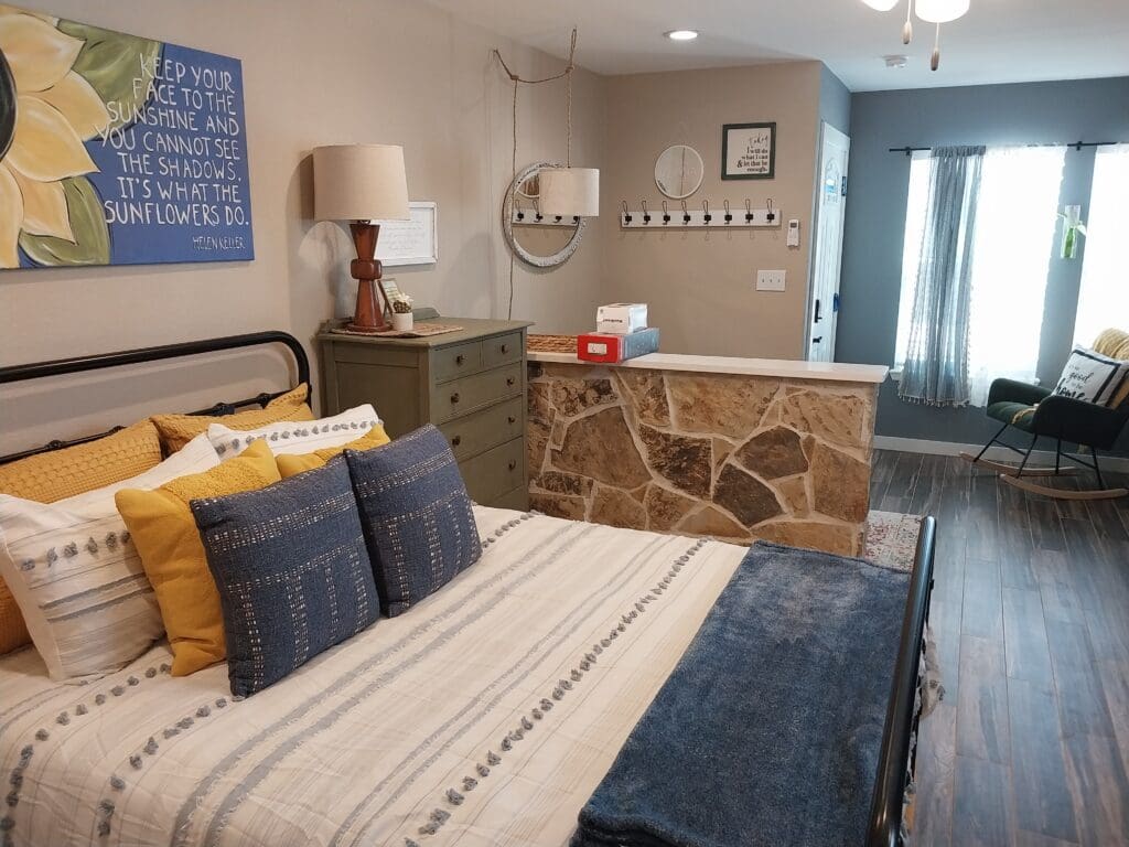 A bedroom with a striped bed, blue and yellow pillows, a wooden dresser, a wall hanging, a stone partition, and a rocking chair near a window with gray curtains.