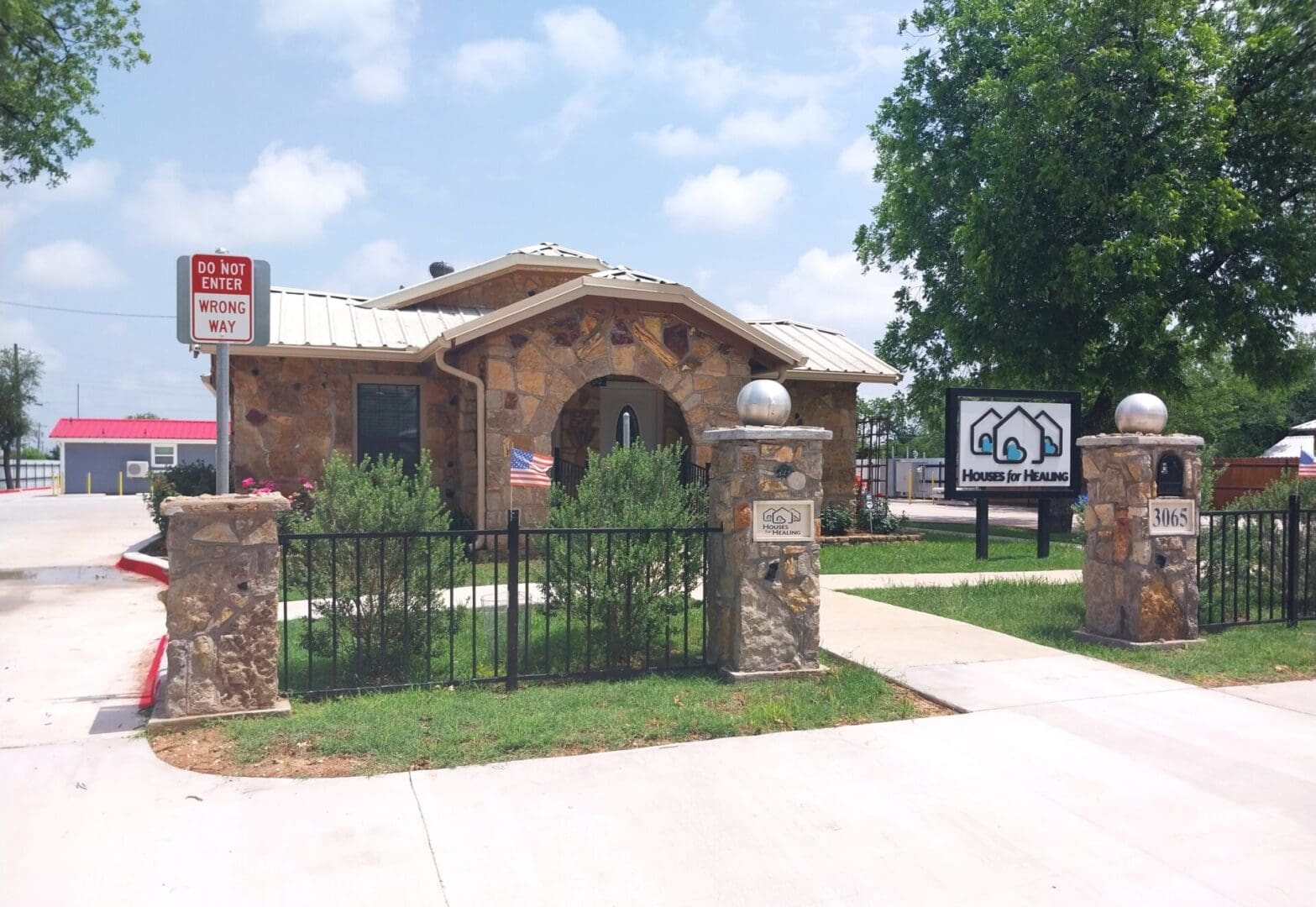 20240503_135317 A small stone building with a metal roof, surrounded by a black fence. A sign reads "Homes by Hhasna." A smaller flag is mounted near the entrance. Trees and blue sky are visible in the background.