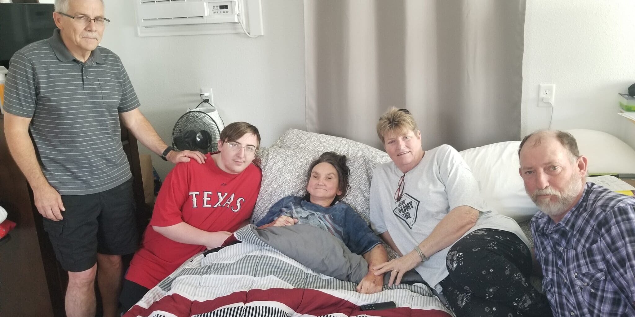 A person lies in bed while surrounded by four people standing and sitting close. The room has an air conditioner above the bed and gray curtains. The bedspread is red, white, and gray striped.