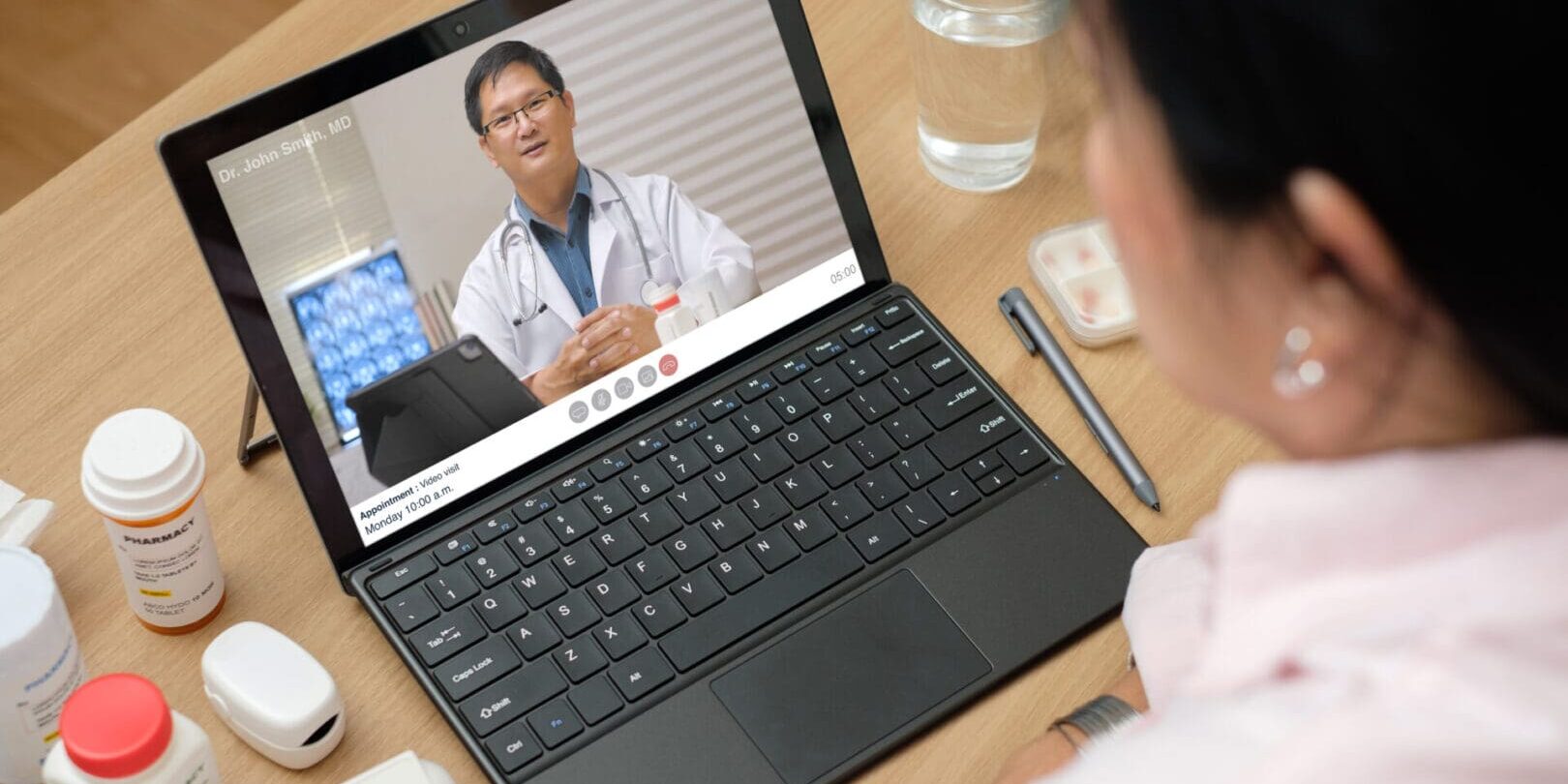 A person is having a video call with a doctor on a laptop. Several medication bottles, a glass of water, and a digital thermometer are on the table around the laptop.